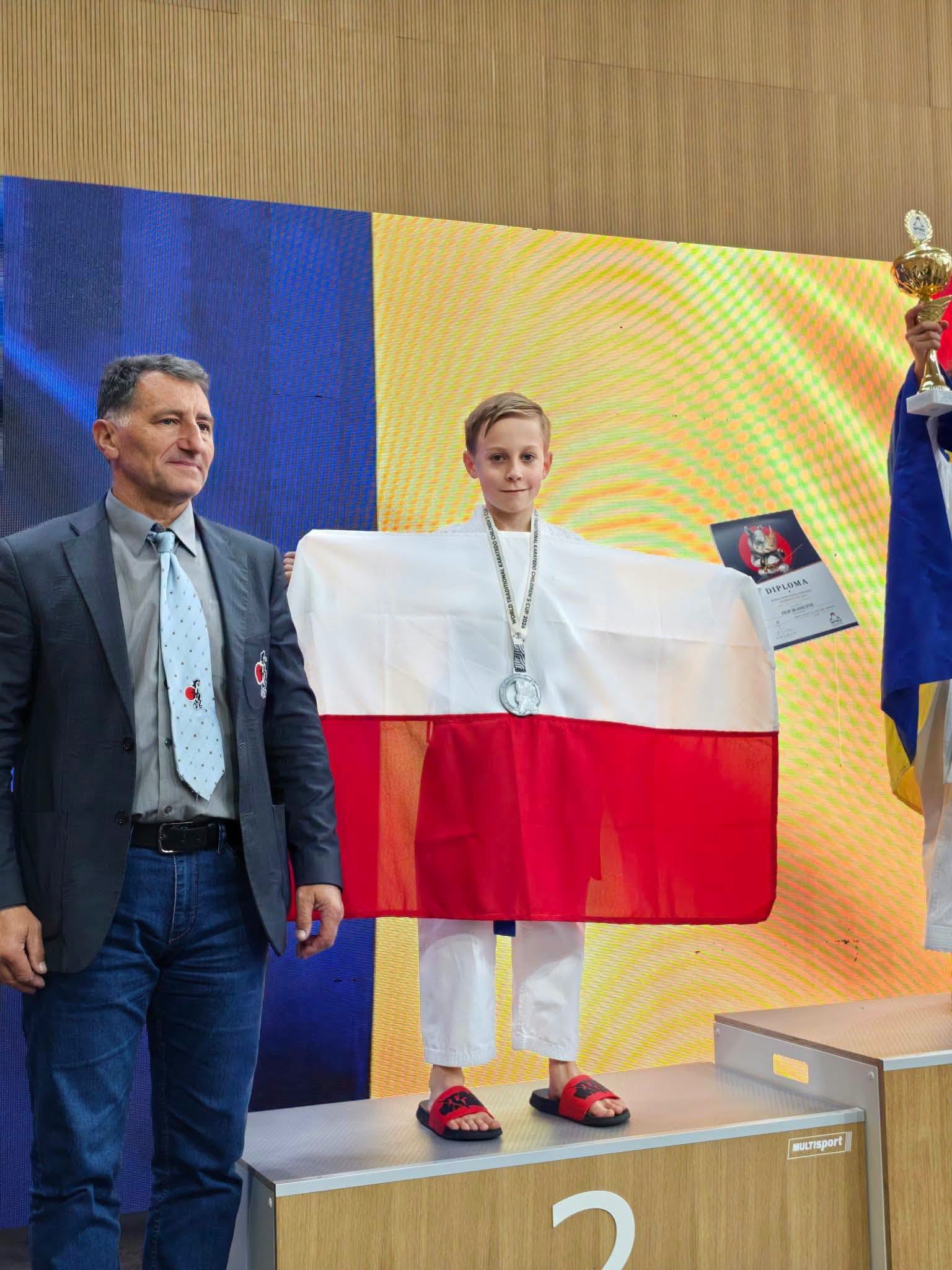 Chłopiec w&nbsp;stroju karate na&nbsp;2 miejscu podium z&nbsp;biało-czerwona flagą i&nbsp;pucharem w&nbsp;dłoni. Obok z&nbsp;lewj strony trener.