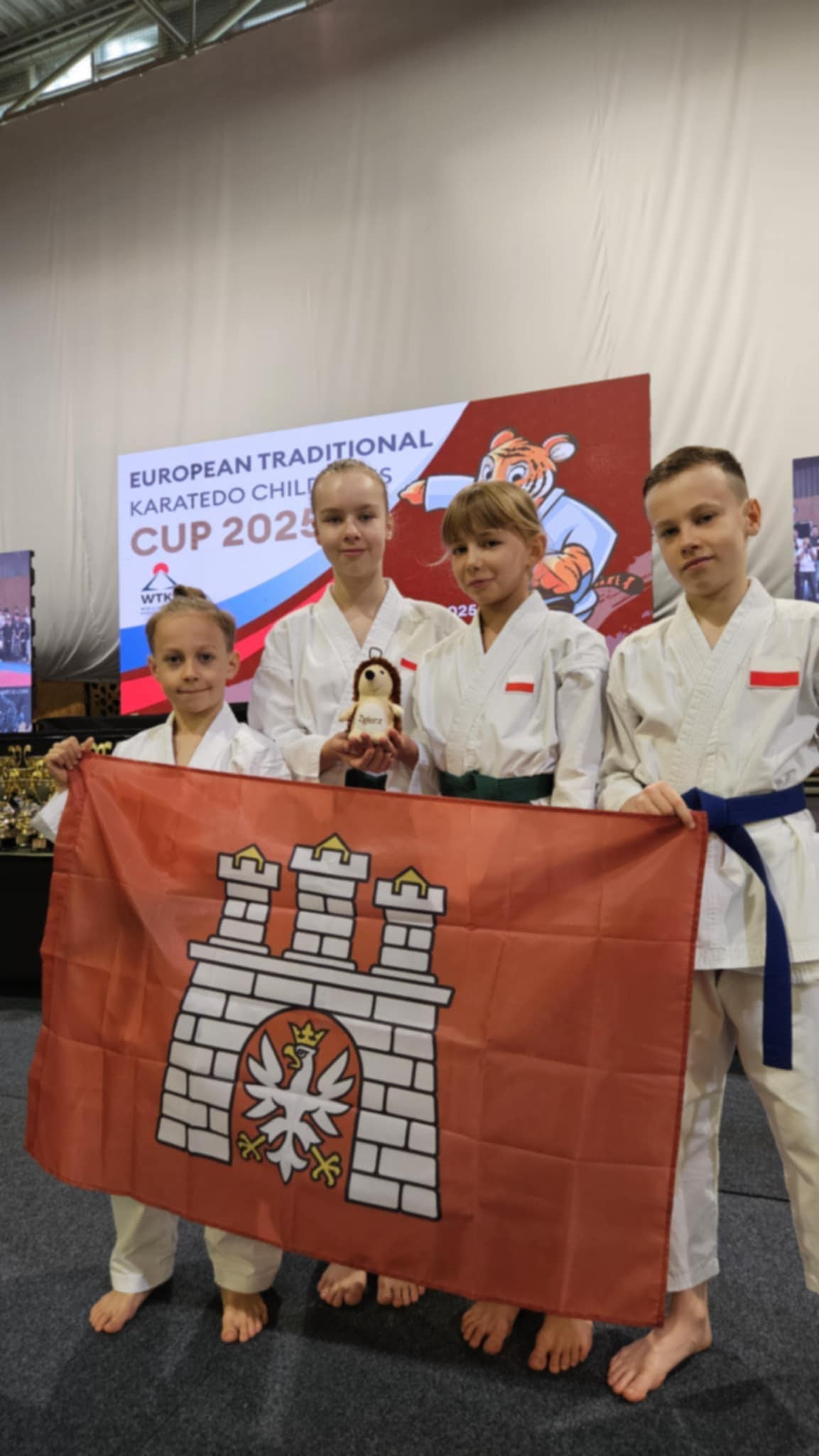 Chłopiec z&nbsp;lewej i&nbsp;3 dziewczynki, w&nbsp;strojach karatek&oacute;w trzymają flagę Zgierza przed zawodami w&nbsp;hali sportowej w&nbsp;Słowenii