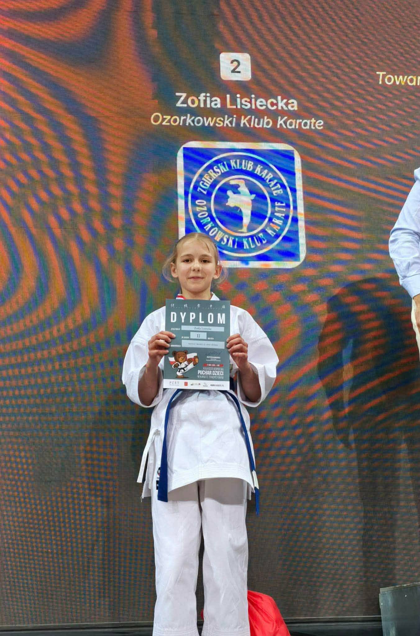 Uczeń w&nbsp;stroju karateki stoi na&nbsp;tle baner&oacute;w z&nbsp;informujących o&nbsp;turnieju i&nbsp;sponsorach pucharu Polski w&nbsp;karate. Trzyma przed sobą dyplom
