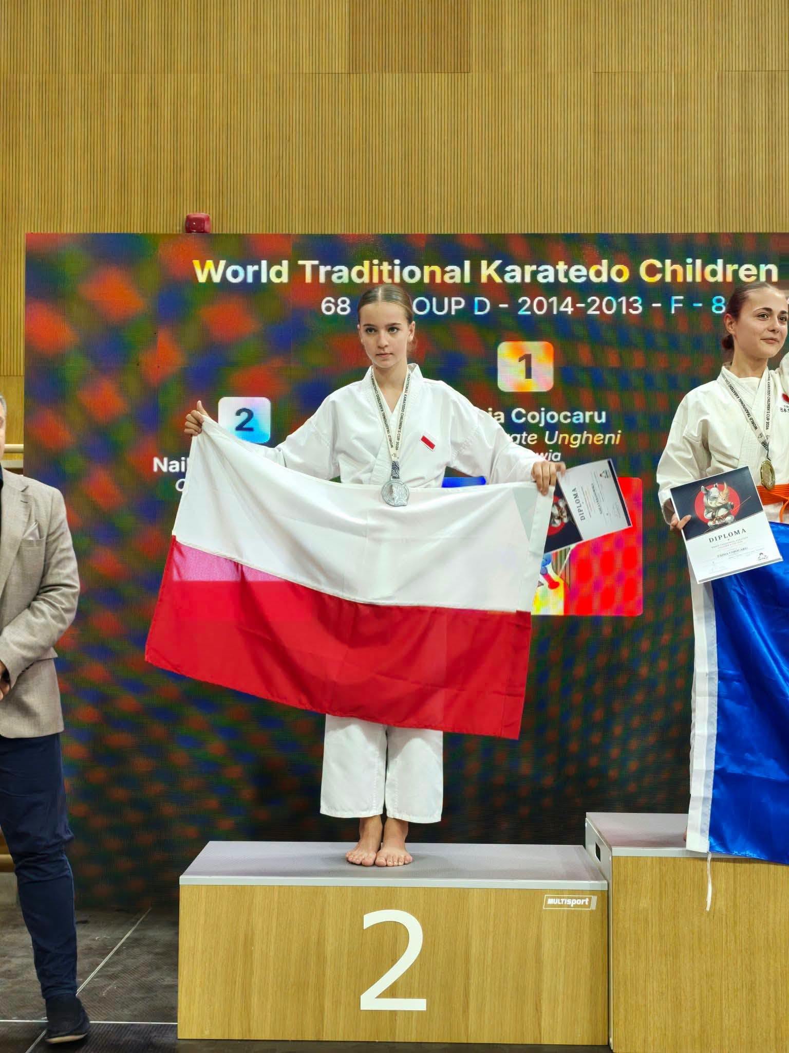 Dziewczynka w&nbsp;stroju karate na&nbsp;2 miejscu podium z&nbsp;biało-czerwona flagą i&nbsp;pucharem w&nbsp;dłoni. Obok z&nbsp;lewj strony trener.