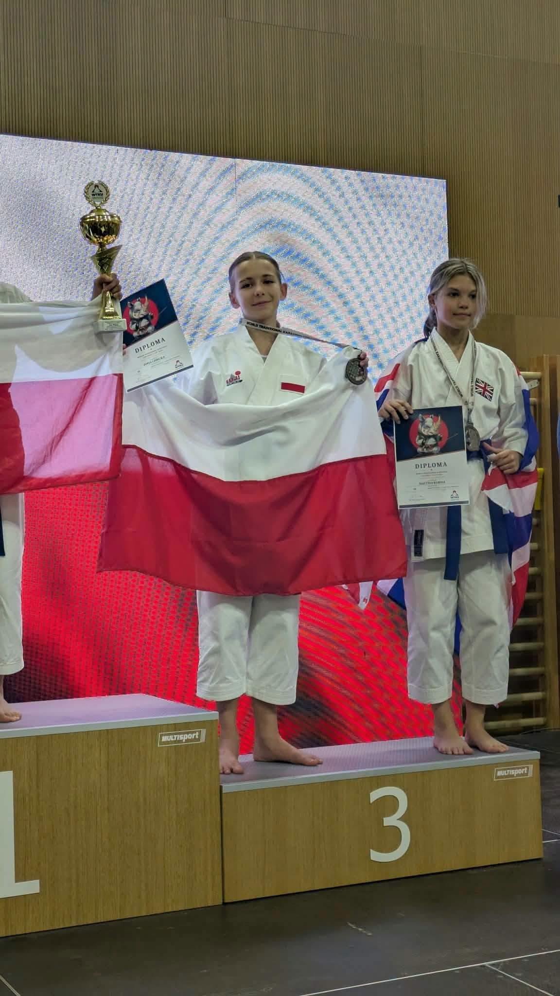Dwie dziewczynki w&nbsp;stroju karate na&nbsp;3 miejscu podium. Jedna z&nbsp;biało-czerwona flagą i&nbsp;pucharem w&nbsp;dłoni. 