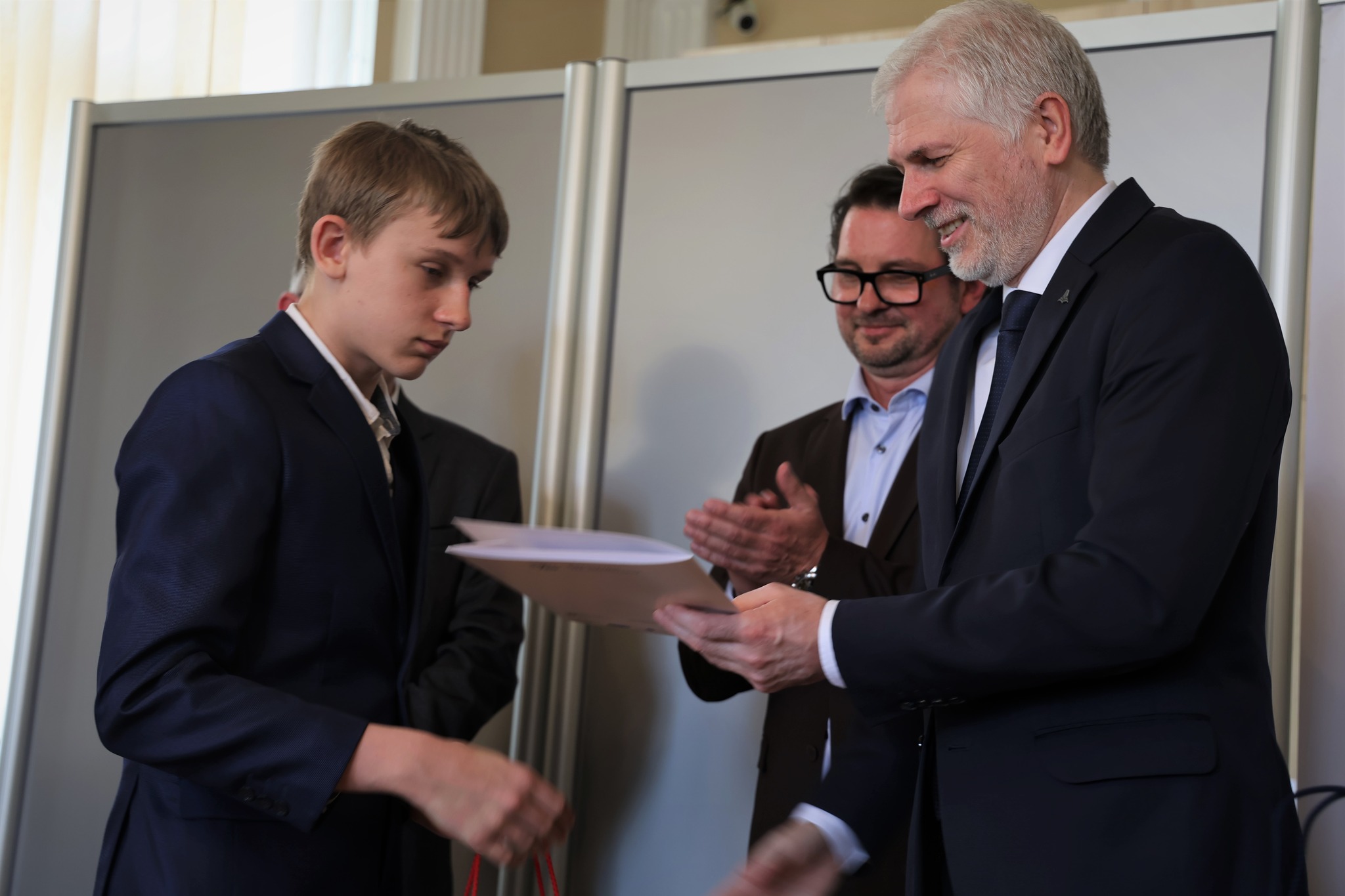 Chłopiec w&nbsp;czarnym garniturze odbiera nagrodę z&nbsp;rąk Kuratora Oświaty