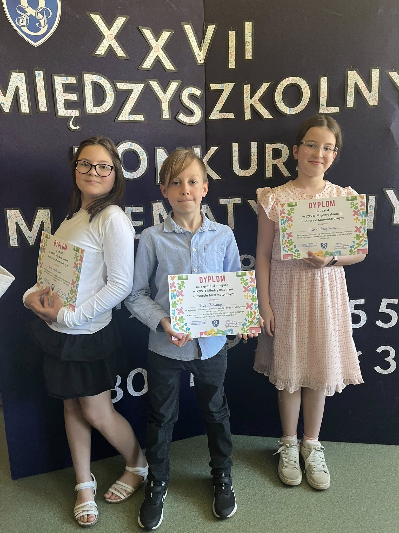 Troje uczni&oacute;w: dwie dziewczynki i&nbsp;chłopiec między nimi , z&nbsp;dyplomami trzymanymi przed soba pozuja do zdjęcia na&nbsp;tel białego napisu na&nbsp;czarnym tle z&nbsp;nazwa konkursu matematycznego
