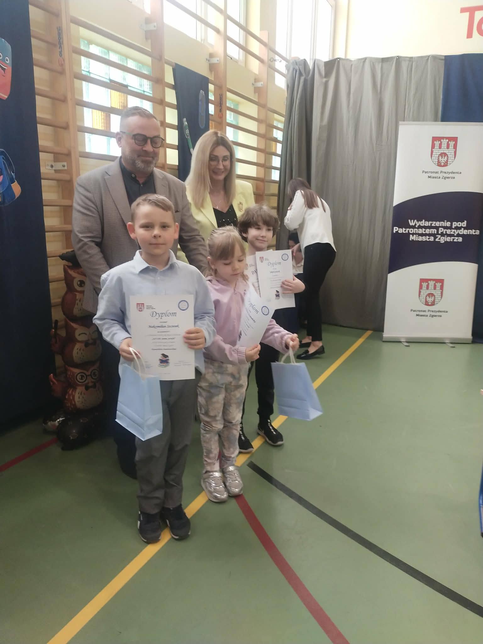 Troje dzieci: dw&oacute;ch chłopc&oacute;w i&nbsp;dziewczynka pozują do zdjęcia na&nbsp;tle ściany z&nbsp;drabinkami w&nbsp;sali gimnastycznej. Za nimi prezydent miasta i&nbsp;dyrektor szkoły