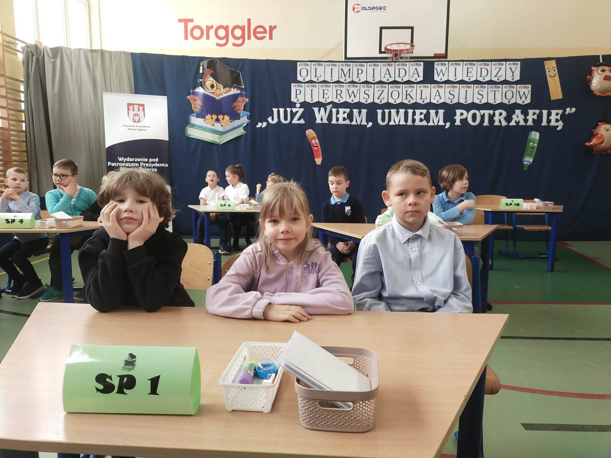 Troje dzieci: dw&oacute;ch chłopc&oacute;w i&nbsp;w środku dziewczynka siedzą przy stoliku ustawionym w&nbsp;sali gimnastycznej z&nbsp;kartką SP1, podczas konkursu. W tle dzieci przy innych stolikach 