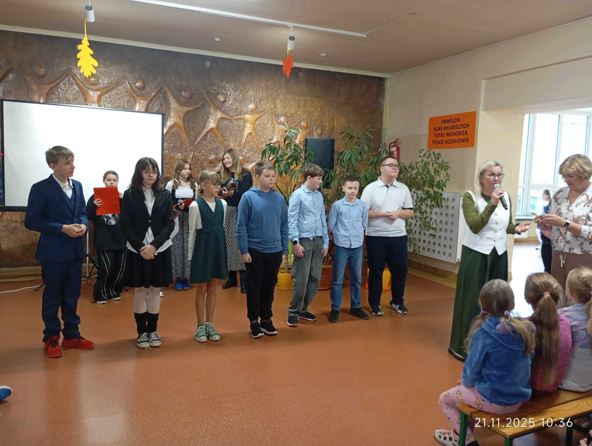 Grupa uczni&oacute;w stoi na&nbsp;szkolnym korytarzu podczas rozdawania nagr&oacute;d w&nbsp;konkursie, przed siedzącą publicznością. W tle Ekran multimedialny i&nbsp;baner z&nbsp;naapisem święto Niepodległości.