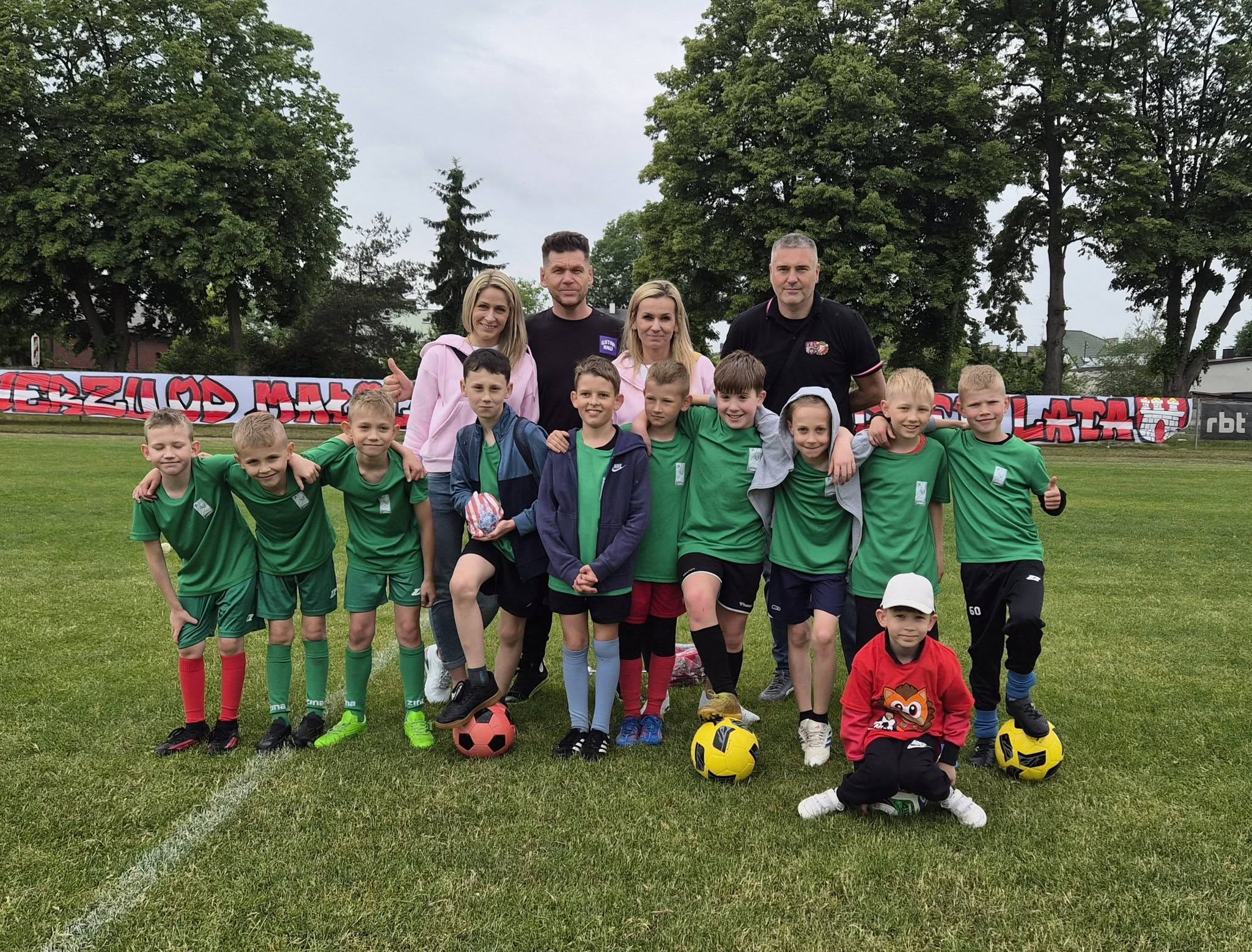 Grpa dzieci - piłkarzy w&nbsp;zielych strojach sportowych wraz z&nbsp;opiekunami pozuje do zdjęcia na&nbsp;murawie stadionu