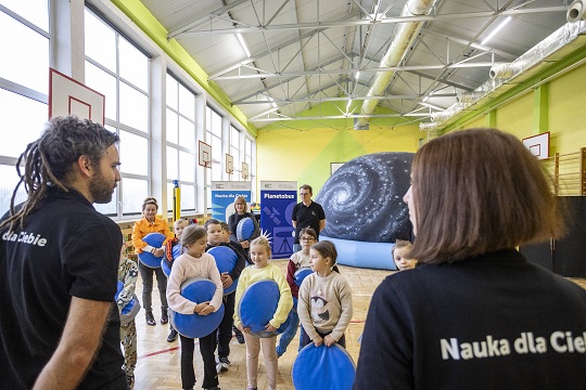 Plakat Centrum Nauki Kopernik - prowadzący, dzieci i&nbsp;kopuła mobilnego planetarium w&nbsp;szkole w&nbsp;sali gimnastycznejuła 