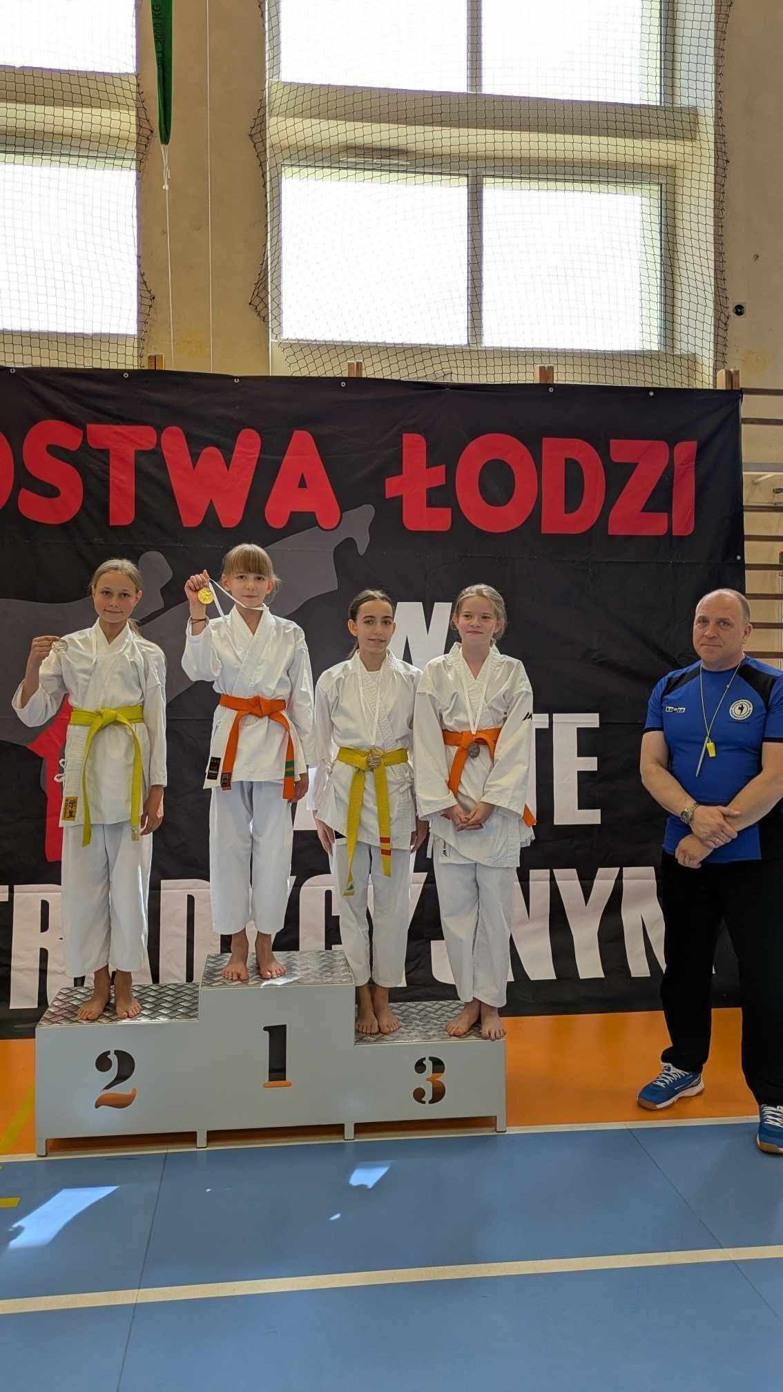 Czterech chłopc&oacute;w stoi na&nbsp;podium w&nbsp;strojach karatek&oacute;w, w&nbsp;tym dw&oacute;ch na&nbsp;3 miejscu. Obok podium po prawej stronie stio trener w&nbsp;niebieskiej koszulce z&nbsp;nadrukiem i&nbsp;czarnych spodniach. W tle baner informujący o&nbsp;Mistrzostwach Łodzi w&nbsp;karate