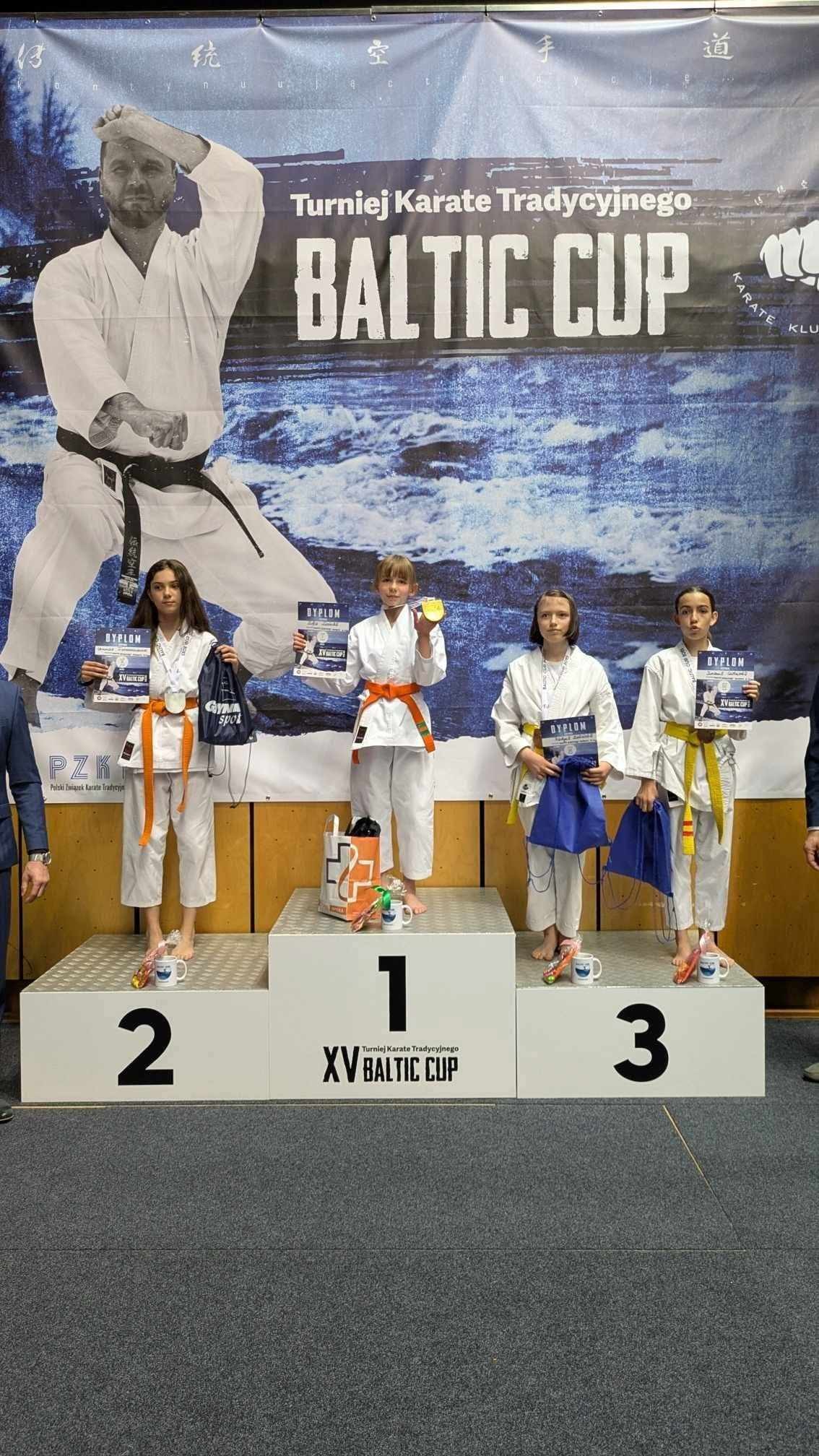 Cztery dziewczyny stoją na&nbsp;podium w&nbsp;strojach karatek&oacute;w, w&nbsp;tym dwie na&nbsp;3 miejscu. Trzymają dyplomy i&nbsp;torby z&nbsp;nagrodami. W tle baner informujący o&nbsp;turnieju karate Baltic Cup