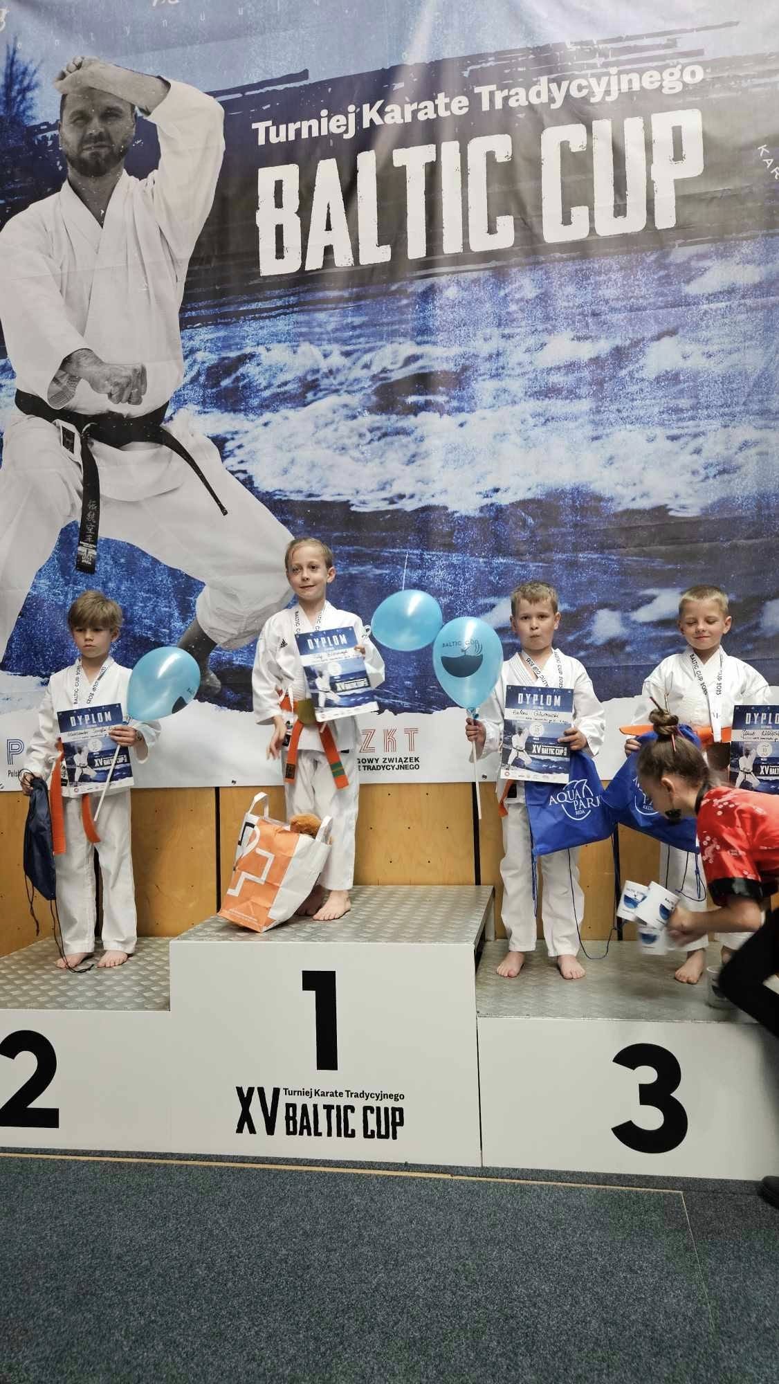 Czterech chłopc&oacute;w stoi na&nbsp;podium w&nbsp;strojach karatek&oacute;w, w&nbsp;tym dw&oacute;ch na&nbsp;3 miejscu. Trzymają dyplomy, niebieskie balony i&nbsp;torby z&nbsp;nagrodami. W tle baner informujący o&nbsp;turnieju karate Baltic Cup