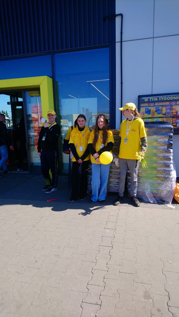 Grupa dzieci w&nbsp;ż&oacute;łtych koszulkach i&nbsp;w ż&oacute;łtych czapkach, trzymająca ż&oacute;łte balony, żonkile i&nbsp;puszki w&nbsp;czasie zbi&oacute;rki w&nbsp;ramach akcji Pola nadziei