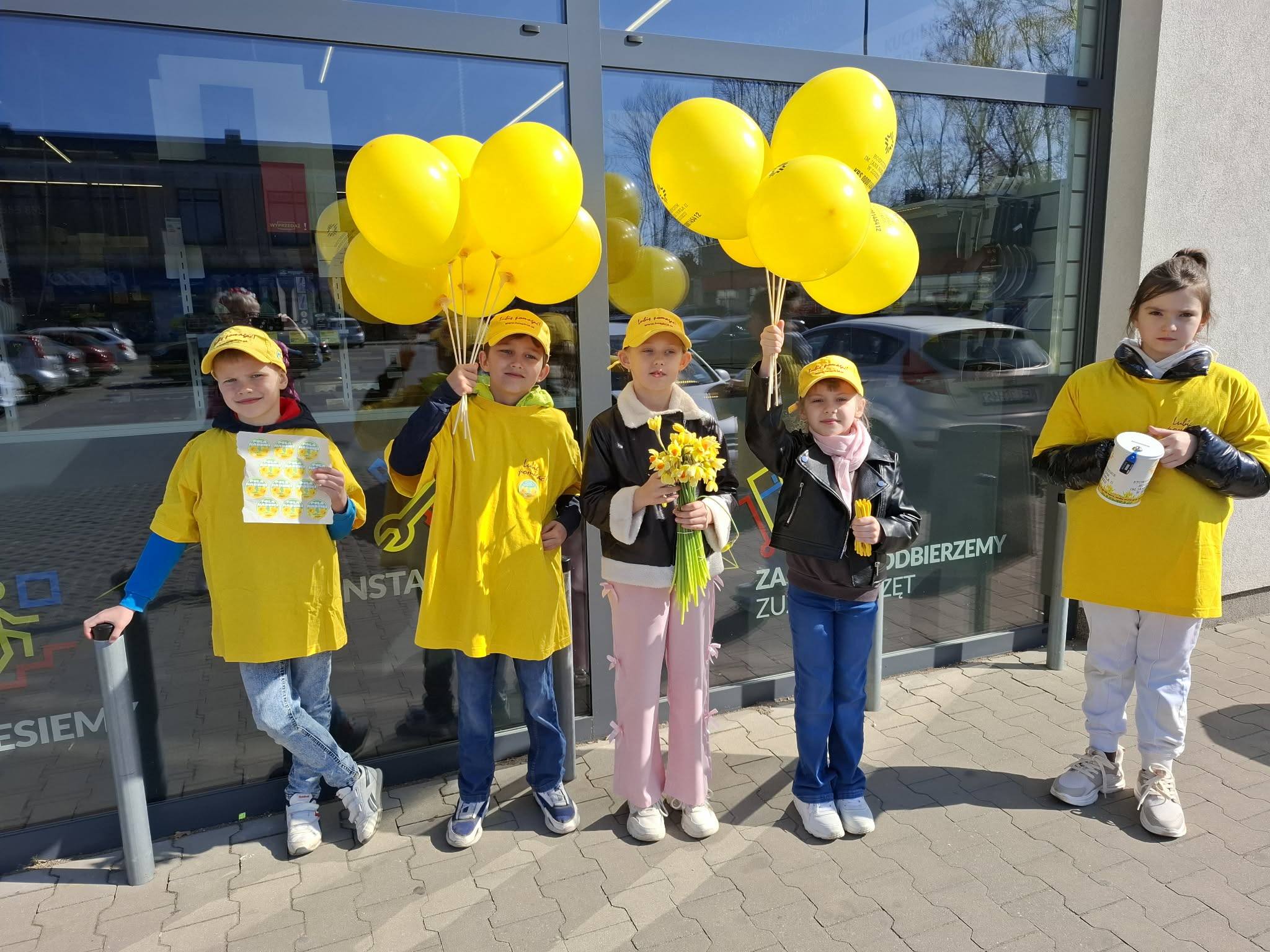 Grupa dzieci w&nbsp;ż&oacute;łtych koszulkach i&nbsp;w ż&oacute;łtych czapkach, trzymająca ż&oacute;łte balony, żonkile i&nbsp;puszki w&nbsp;czasie zbi&oacute;rki w&nbsp;ramach akcji Pola nadziei