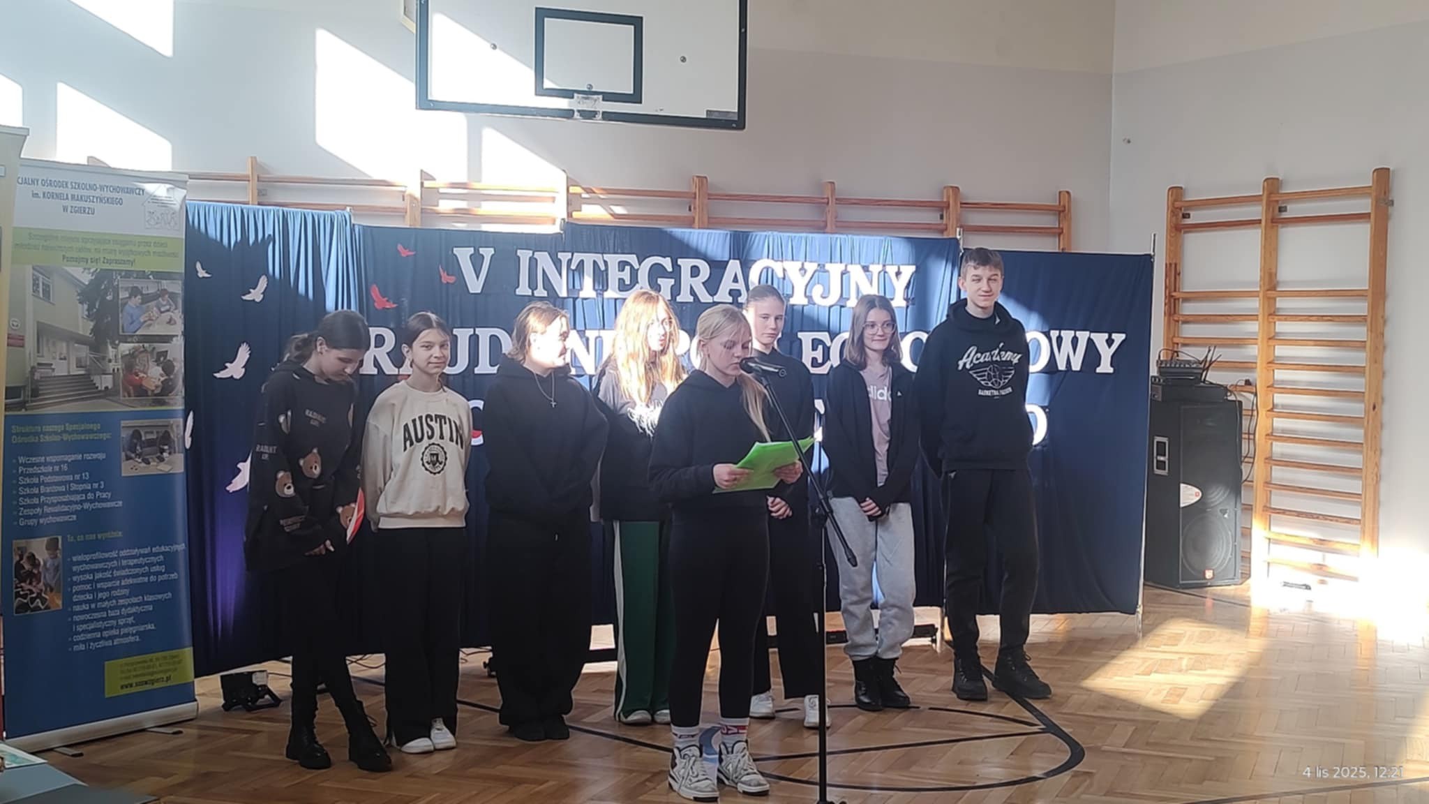 Grupa uczni&oacute;w prezentuje wykonanie zadań podczas V Integracyjnego raju Niepodległościowego. Stoją na&nbsp;środku sali gimnastycznej na&nbsp;tle baneru z&nbsp;napisem informującym o&nbsp;rajdzie 