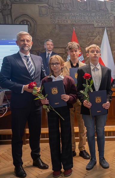 Dwoje uczni&oacute;w pozuje do zdjęcia z&nbsp;prezydentem miasta, trzymając przed soba czerwone r&oacute;że i&nbsp;dyplomy