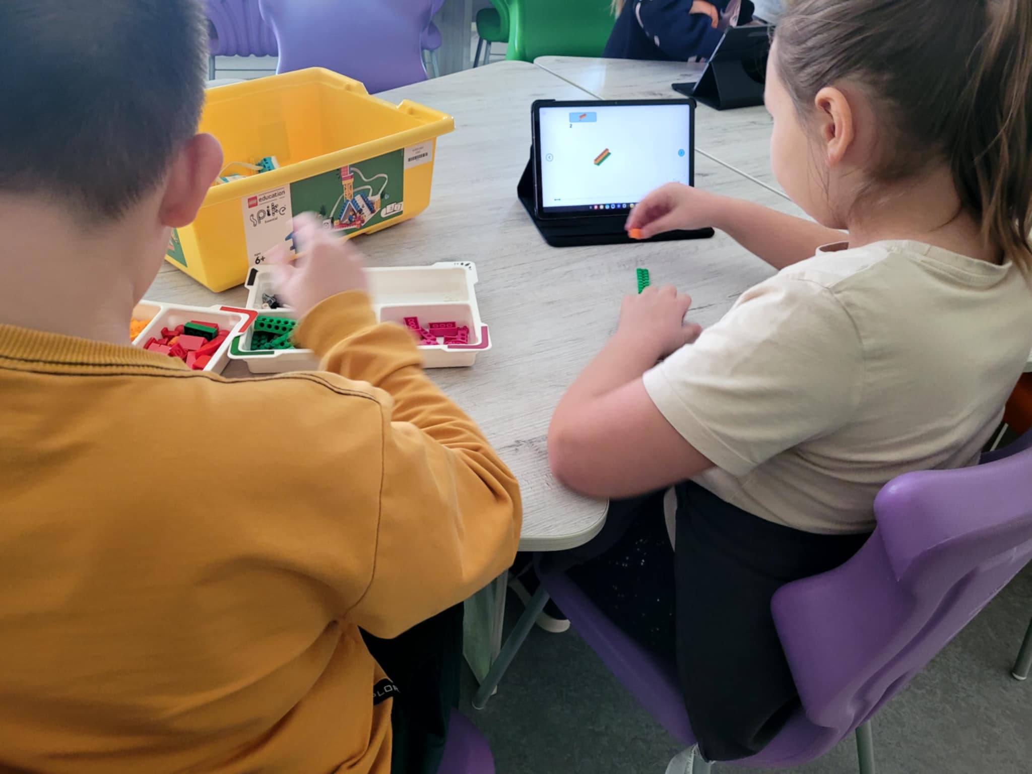 Dziewczynka i&nbsp;chłopiec siedzą przy stole z&nbsp;uruchomionymi tabletami i&nbsp;klockami Lego