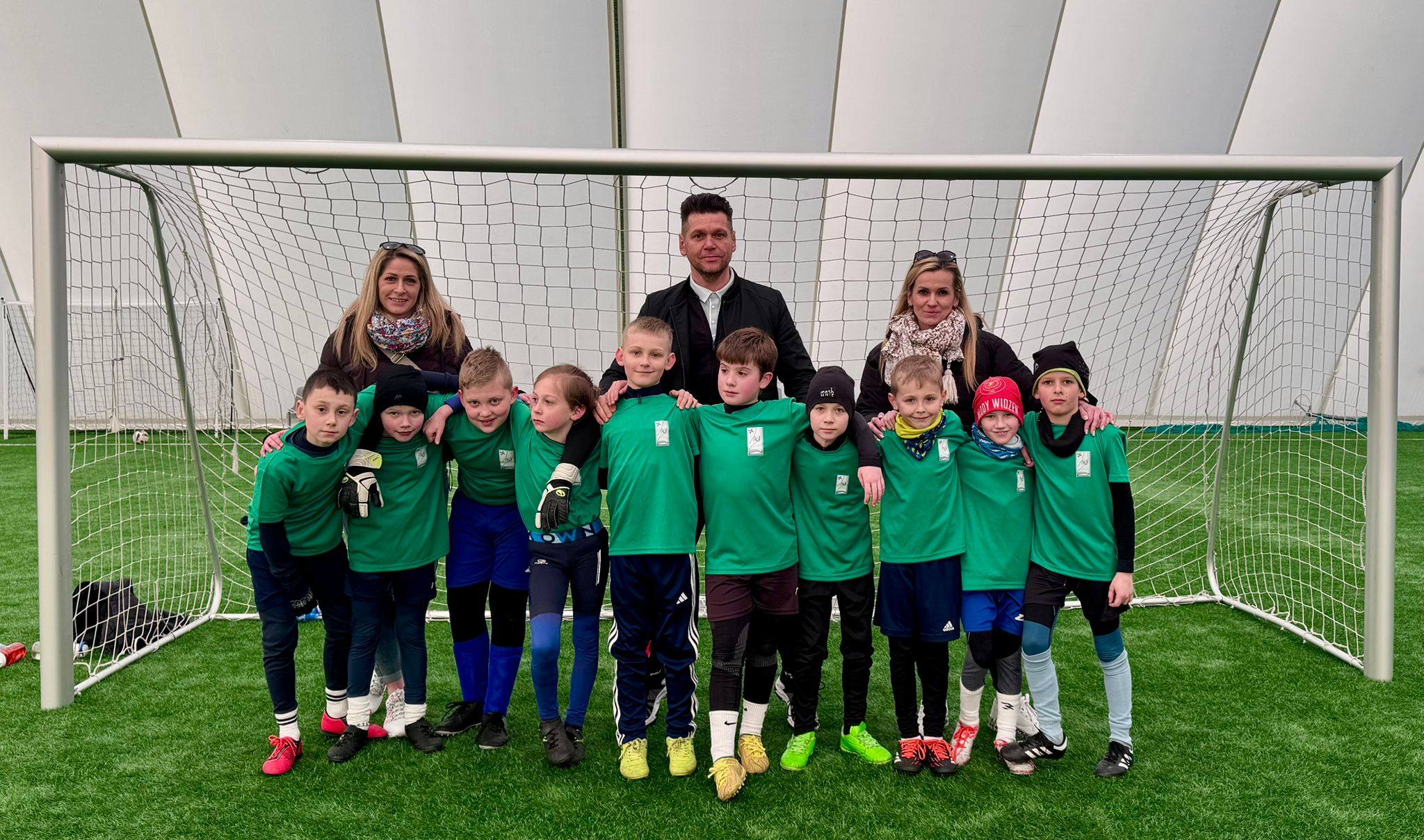 Grupa uczni&oacute;w z&nbsp;opiekunami w&nbsp;zielonych koszulkach i&nbsp;białych spodenkach stująca w&nbsp;bramce na&nbsp;boisku sportowym pod kopułą