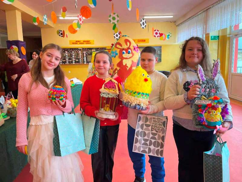 Czworo uczni&oacute;w - 3 dziewczynki i&nbsp;1 chłopiec -stoi na&nbsp;szkolnym korytarzu. Trzymają przed sobą wykonane na&nbsp;konkurs pisanki i&nbsp;prace związane z&nbsp;wielkanocą oraz&nbsp;torby z&nbsp;nagrodami.