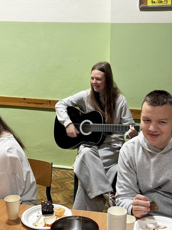 Grupa uczni&oacute;w przy nakrytym stole podczas wigili koła chemicznego i&nbsp;dziewczyka w&nbsp;szarej bluzie siedząca po prawej stronie z&nbsp;gitarą