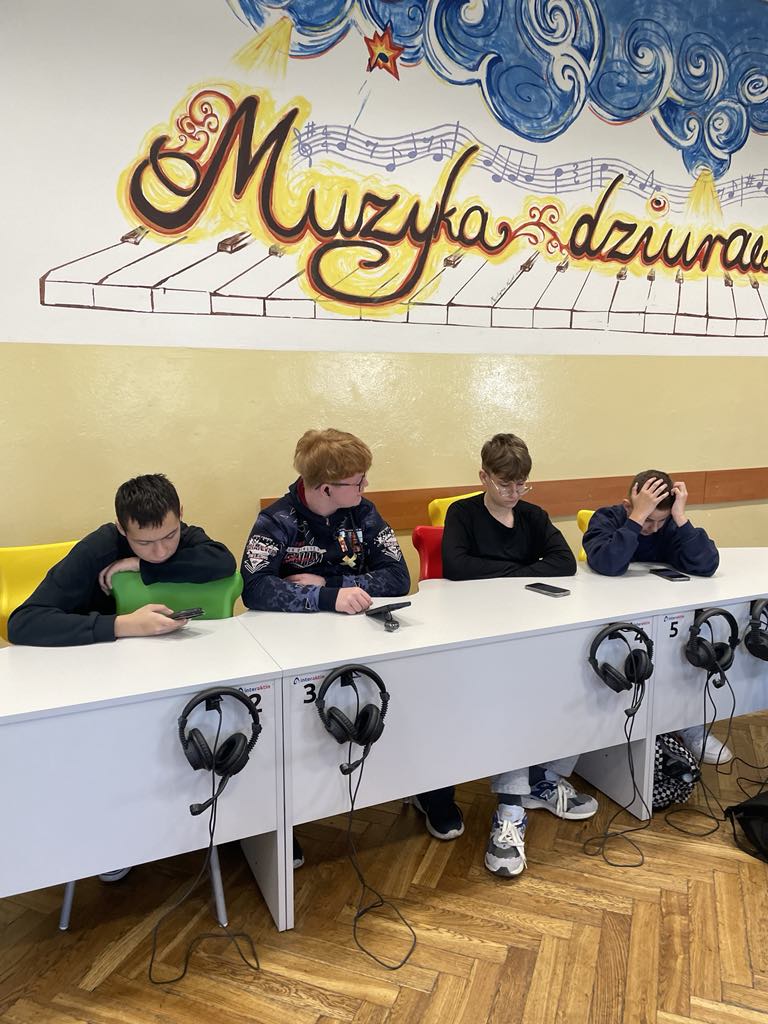 Grupa 4 uczni&oacute;w w&nbsp;czrnych bluzach przy jasnoszarych stolikach w&nbsp;pracowni językowej. Przy każdym stoliku wiszą czarne słuchawki. Uczniowie wykonują polecenia na&nbsp;telefonach podczas warsztat&oacute;w międzynarodowych w&nbsp;języku angielskim.