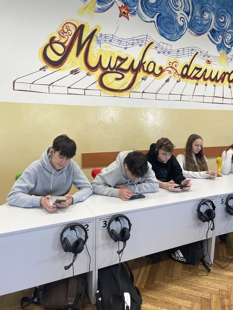 Grupa 4 uczni&oacute;w przy jasnoszarych stolikach w&nbsp;pracowni językowej. Przy każdym stoliku wiszą czarne słuchawki. Uczniowie wykonują polecenia na&nbsp;telefonach podczas warsztat&oacute;w międzynarodowych w&nbsp;języku angielskim.