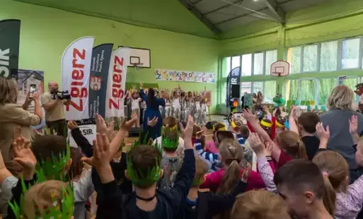 Duża grupa dzieci z opiekunami na sali gimnastycznej. Dzieci stoją z podniesionymi rękami. Niektóre mają zielone korony z rysunkami jeża na głowach. Wykonując gesty wskazywane przez grupę uczniów występujących w srebrnych strojach przed nimi. Na ścianie n