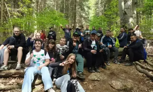 Duża grupa uczniów odpoczywajaca w środku lasu, na szlaku, w czasie wędórwki na górę. Wszyscy siedzą na zwalonym pniu drzewa lub kamieniach przy szlaku.
