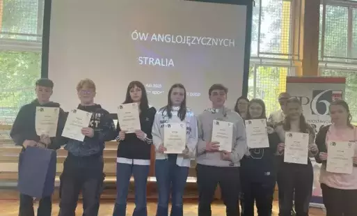 Grupa 8 uczniów z dyplomami w dłoniach stoi w sali gimnastycznej na tle ekranu z wyświetloną nazwą imprezy: IX Festiwal Krajów Anglojęzycznych - Australia. Za uczniami stoi naczelnik wydziału edukacji i pani organizatorka imprezy. Po prawej stronie za ucz