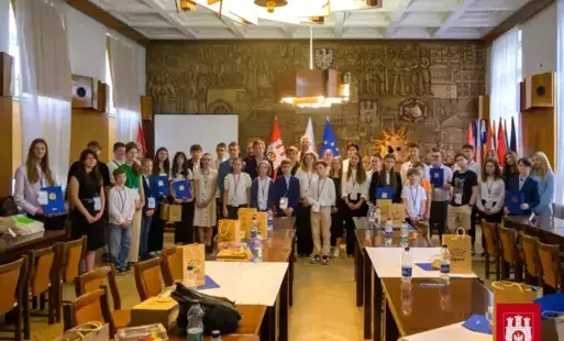 Grupa uczniów zgieraskich szkół z prezydentem miasta i innymi pracowni stoi w sali obrad Urzedu Miasta Zgierza. Uczniowie mają zawieszone identyfikatory i rzymają przed sobą certfikaty.