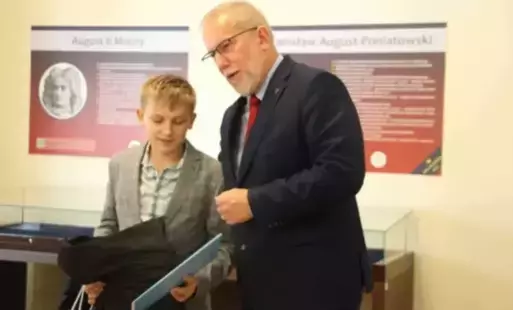 Chłopiec w szarej marynarce i koszuli w kratkę odbiera nagrodę z rąk dyrektora Archiwum Państwowego. W tle na beżowej ścianie umieszczone tablice historyczne