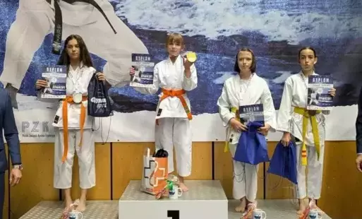 Na podium w strojach karateków stoją 4 dziewczyny, w tym dwie na podium na miejscu trzecim. Wszystkie trzymają dyplomy i torby z nagrodami. Podium ustawione jest na tle baneru reklamującego zawody Baltic Cup: karateka i napisy