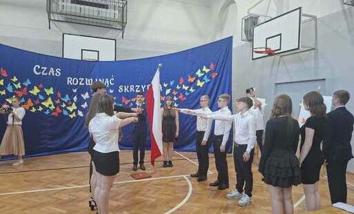 Grupa uczniów ósmych klas składa ślubowanie na sali gimnastycznej. Stoją w galowych strojach po dwóch stronach flagi państwoej z podniesionymi w jej kierunku prawymi rękami. W tle granatowa dekoracja: na materiale kolorowe motyle