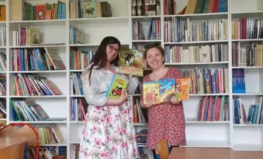 Dwie panie bibliotekarki stoją w bibliotece na tle półek z książkami: od lewej wyższa w sukience w kwiaty i jensowej kurtce, po prawej niższa w beżowej sukience, prezentują zakupione nowe książki, które trzymają przed sobą.