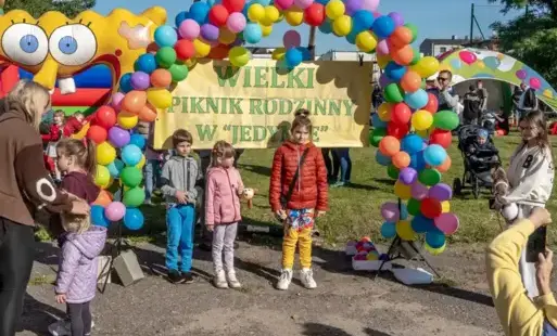 W wielkim łuku z kolorowych balonów stojącym na szkolnym boisku wisi napis Wielki piknik rodzinny i stoi trójka dzieci pozująca do zdjęć: od lewej chłopiec w szarej kurtce i niebieskich spodniach, w środku dziewczynka w różowej kurtce i szarych leginsach,