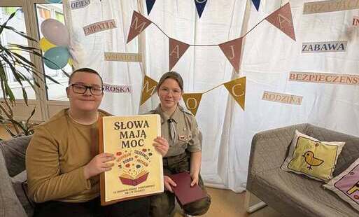Chłopiec siedzący przed tablicą z hasłami dotyczącymi bezpieczeństwa, życzliwości i tolerancji, trzymający plakat z napisem Słowa mają moc. Za nim stoi dziewczynka w stroju harcerki.