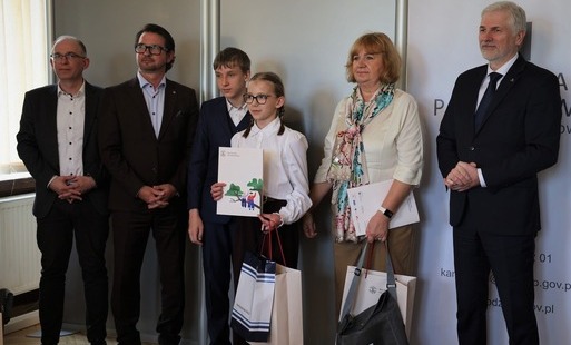 Dwoje uczniów w strojach galowych pozuje do zdjęcia po odbiorze nagród w konkursie. Od lewej stoi dwóch panów w garniturach; w środku chłopiec i dziewczynka; od prawej nauczyciel i kurator oświaty