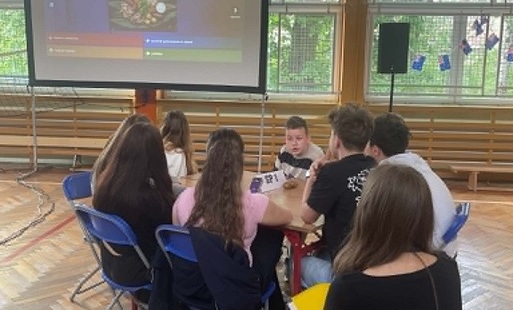 Grupa uczniów siedzi przy stoliku ustawionym w sali gimnastycznej na tle monitora z wyświetloną nazwą imprezy - Festiwal krajów anglojęzycznych