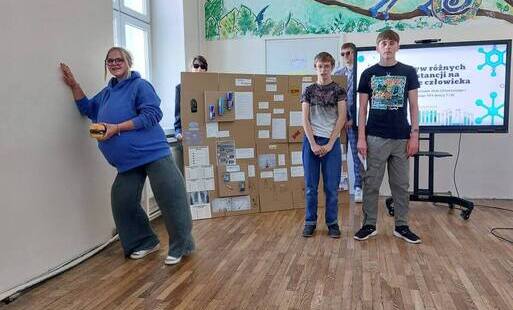 Troje uczniów podczas prezentacji projektu edukacyjnego. Dwóch chłopców na tle monitora z wyświetlonym tytułem projektu prezentuje projekt. Dziewczynka oparta ręką o ścianę przedstawia zachowanie kobiety w ciązy pod wpływem środków odurzających.