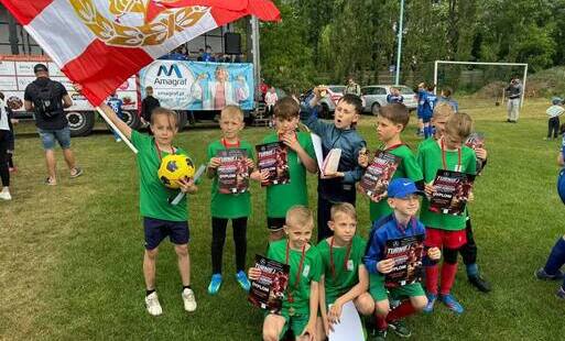 Grupa dzieci - piłkarzy w zielonych strojach sportowych i dyplomami w ręku pozuje do zdjęcia na murawie stadionu