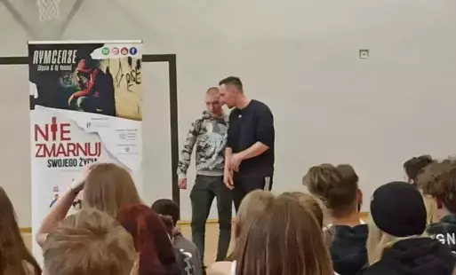 Przed kolorowym banerem z napisem 'Nie zmarnuj swojego życia' stoli w czarnej koszulce muzyk raper z opuszczonym mikrofonem w prawej ręce. Obok stoi uczeń w szarej bluzie. Na pierwszym planie widoczne od tyłu głowy grupy uczniów siedzących i oglądających 