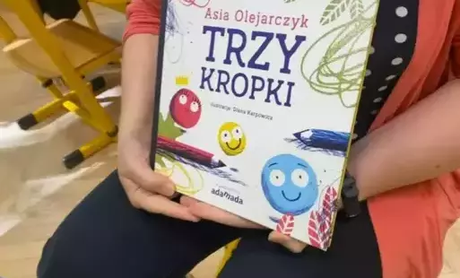 Dziewczynka wczarnych spodniach, czrnej bluzce w białe kropki i pomarańczowym sweterku, siedzi na krześle (nie widać głowy), trzyma w dłoniach swoją autorską ksążkę Trzy kropki. 