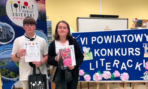 Dwoje uczniów pozuje do zdjęcia z dyplomami i nagrodami otrzymanymi w konkursie. Stoją na tle banerów prezentujących konkur i sponsorów.