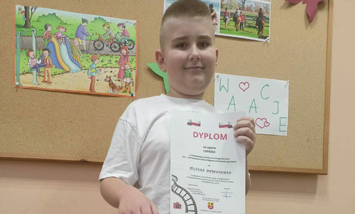 Chłopiec w galowym stroju z dyplomem i torebką z nagrodami pozuje do zdjęcia na tle tablicy z plakatami.